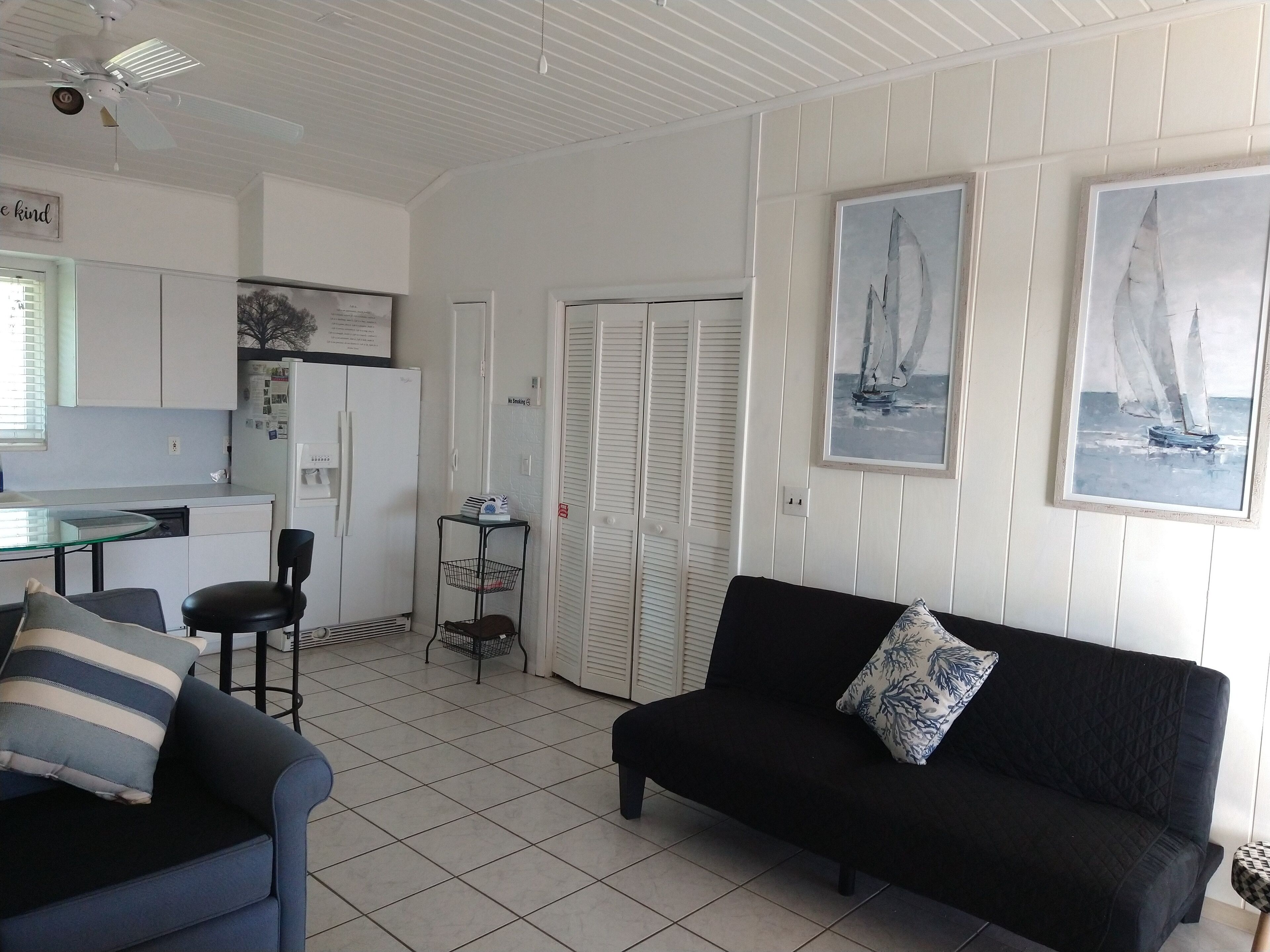 Ein Wohnzimmer mit einer schwarzen Couch, einem blauen Sessel und einem Glastisch. Es gibt eine Küchenzeile mit einem Kühlschrank, einer Mikrowelle und einer kleinen Kochinsel mit Spüle.