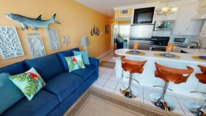 A modern living room with a blue sofa, tropical-themed cushions, and a kitchen area with white cabinets and a central island. - Sunny Poolside Retreat | Bright & Stylish Condo with Resort Amenities! (Galveston)
