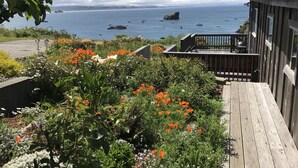 A wooden deck with a barbecue grill, overlooking a coastal area with boats and a rocky outcrop.