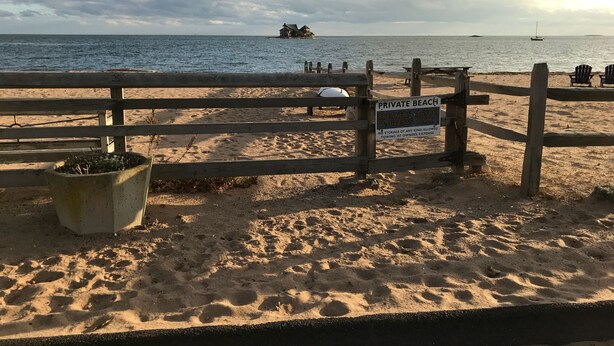 Una playa con una cerca de madera, un letrero que indica que es una playa privada para los miembros de la asociación de surfistas y huéspedes solamente, y un barco a lo lejos.