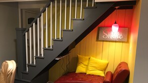 Una escalera con acabado oscuro y barandillas blancas que conduce a un nivel inferior. Un sofá de cuero rojo con cojines amarillos está ubicado frente a una pared amarilla, iluminado por un aplique rojo.