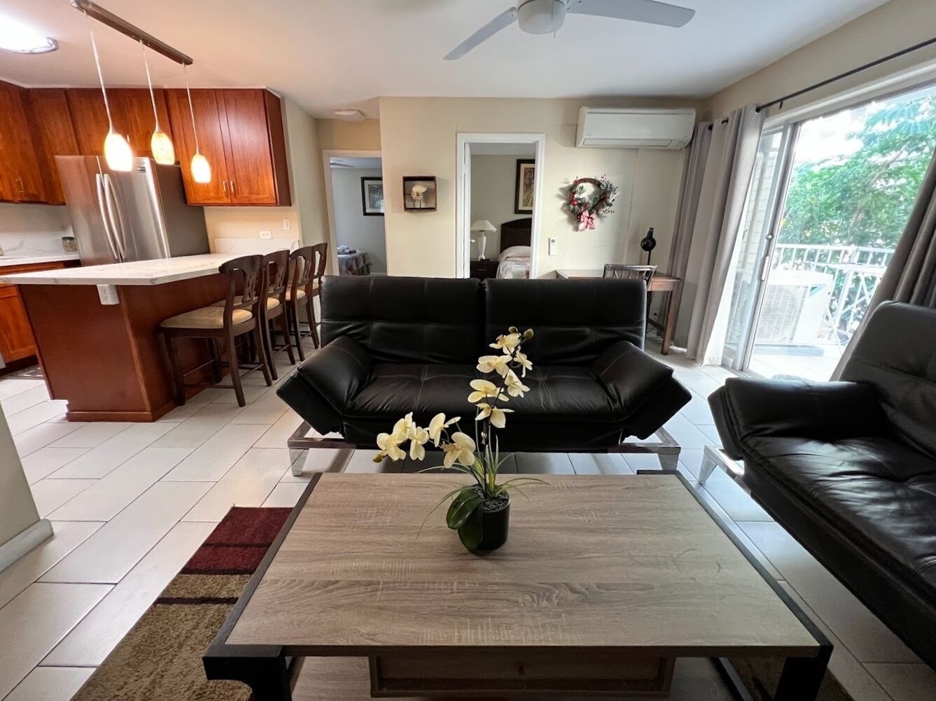 A modern living room with a black leather sofa, a wooden coffee table with a flower vase, and a kitchen area in the background.
