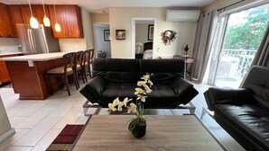 Ett modernt vardagsrum med en svart lädersoffa, ett träbord med en blomvas och ett köksområde i bakgrunden.
