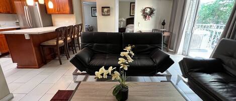 A modern living room with a black leather sofa, a wooden coffee table with a flower vase, and a kitchen area in the background.