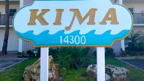 A sign with the name "KIMA" and the number "14300" in front of a building with balconies and palm trees.