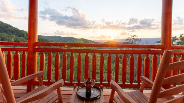 A wooden deck with a table set for two, overlooking a mountainous landscape during sunset. - A Heavenly View ~ 6 BR Indoor Pool Cabin ~ Sleeps 28 ~ Incredible Views! (Sevierville)