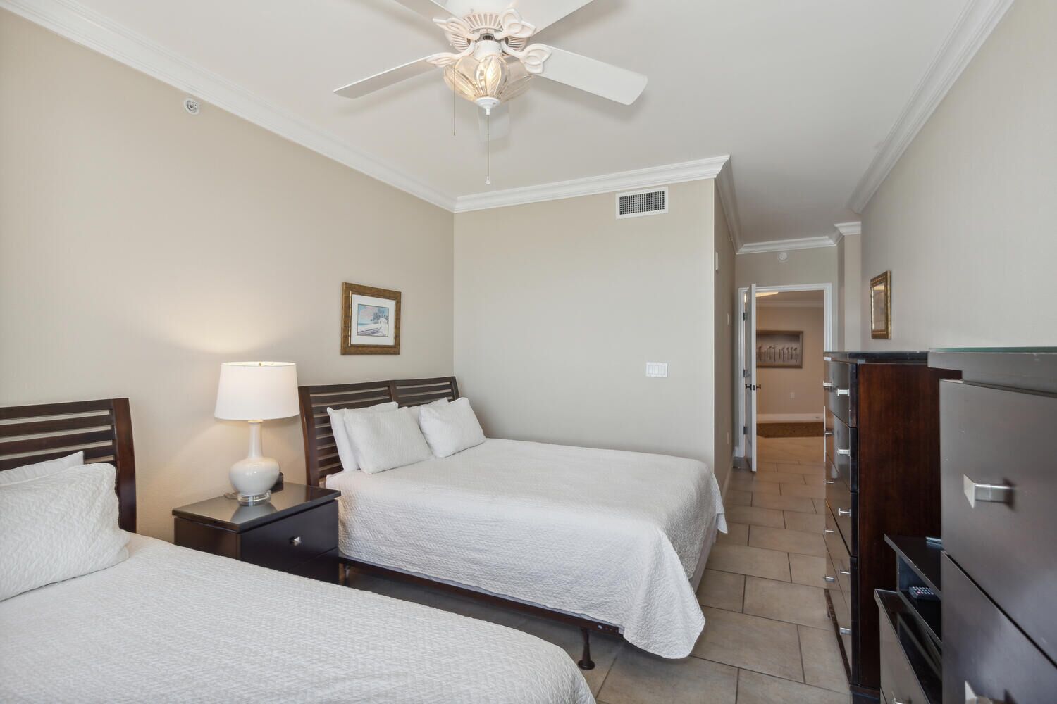 Une chambre d’hôtel avec deux lits, un ventilateur de plafond, une table de chevet avec une lampe et une commode.