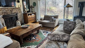 Un salon avec une cheminée en pierre, un téléviseur, un canapé, une table basse et une grande fenêtre donnant sur les arbres.