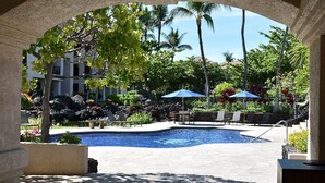 Un área de piscina con sillones de descanso y sombrillas, rodeada de plantas tropicales y palmeras.