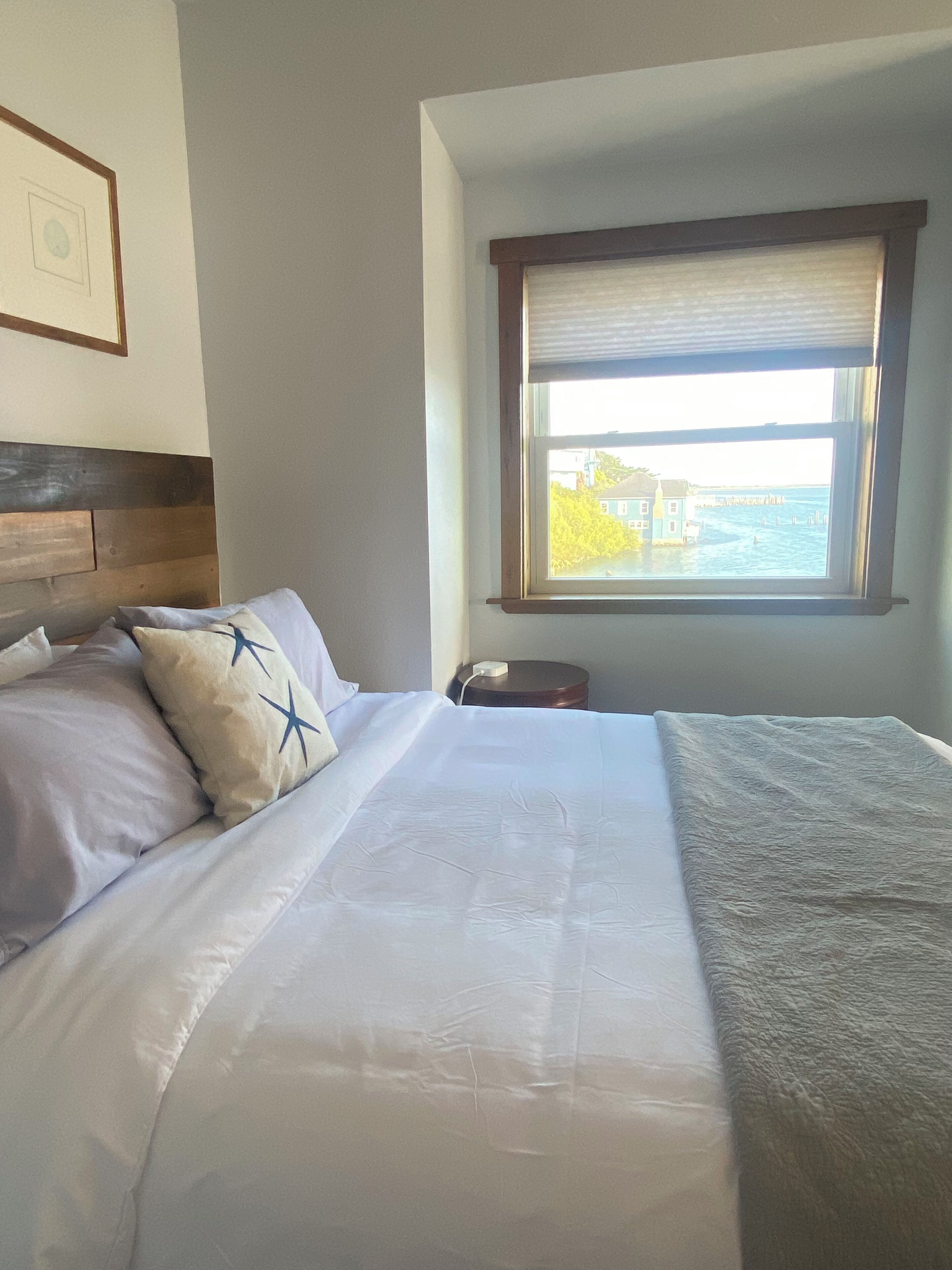 Cama bem arrumada com lençóis brancos e manta cinza, mesa de cabeceira com telefone e janela com vista para um corpo de água e edifícios.