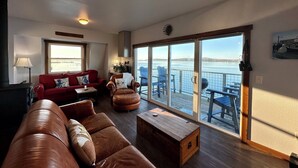 Amplia sala de estar con un sofá rojo, un sofá de cuero marrón y una mesa de centro de madera. Un gran ventanal corredera da a una terraza exterior con sillas azules y vistas al agua.