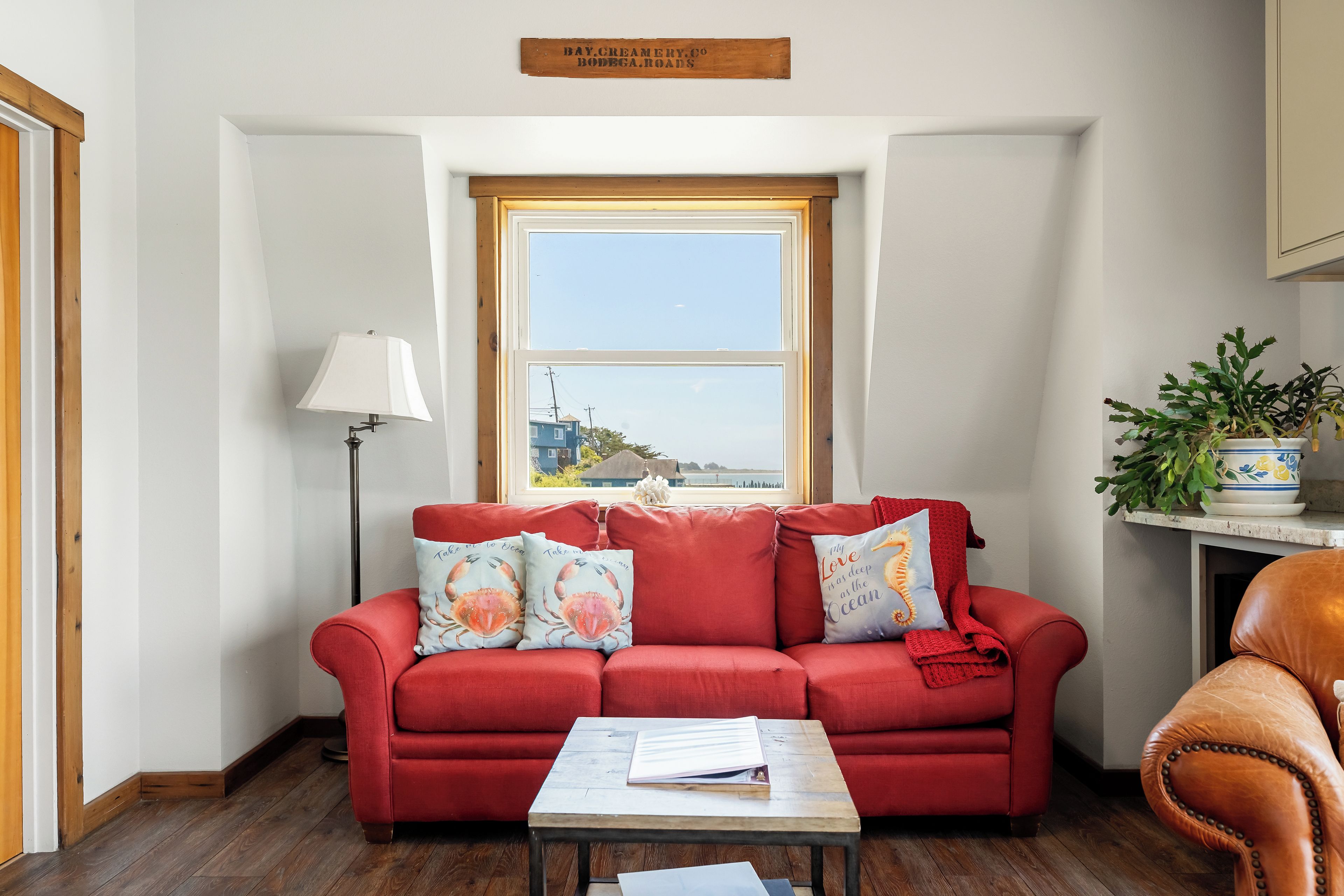A red sofa with ocean-themed cushions, a small table with a book, and a view of the sea through a window.