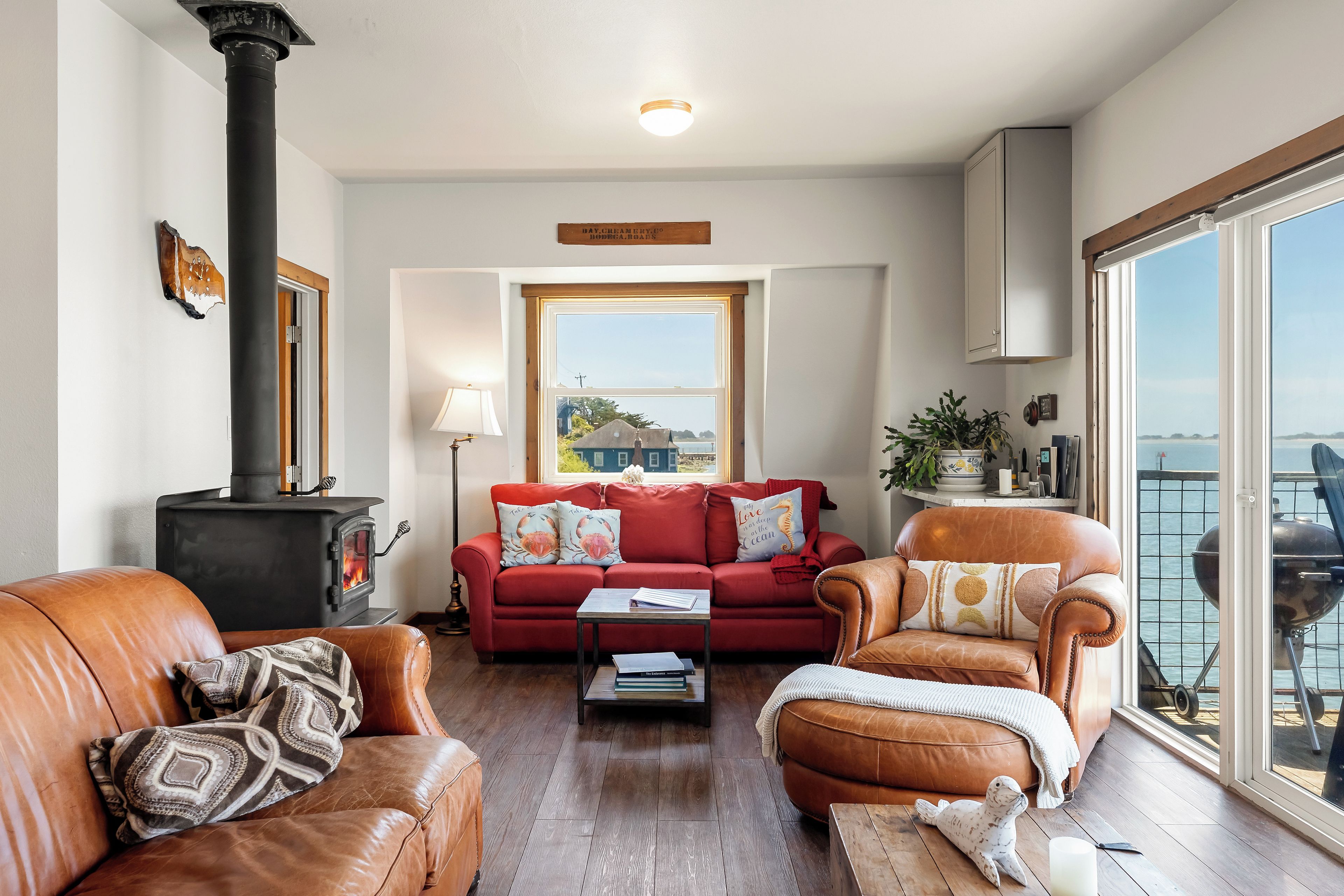 A living room with a fireplace, a sofa, a red couch, and a view of the sea through large windows.