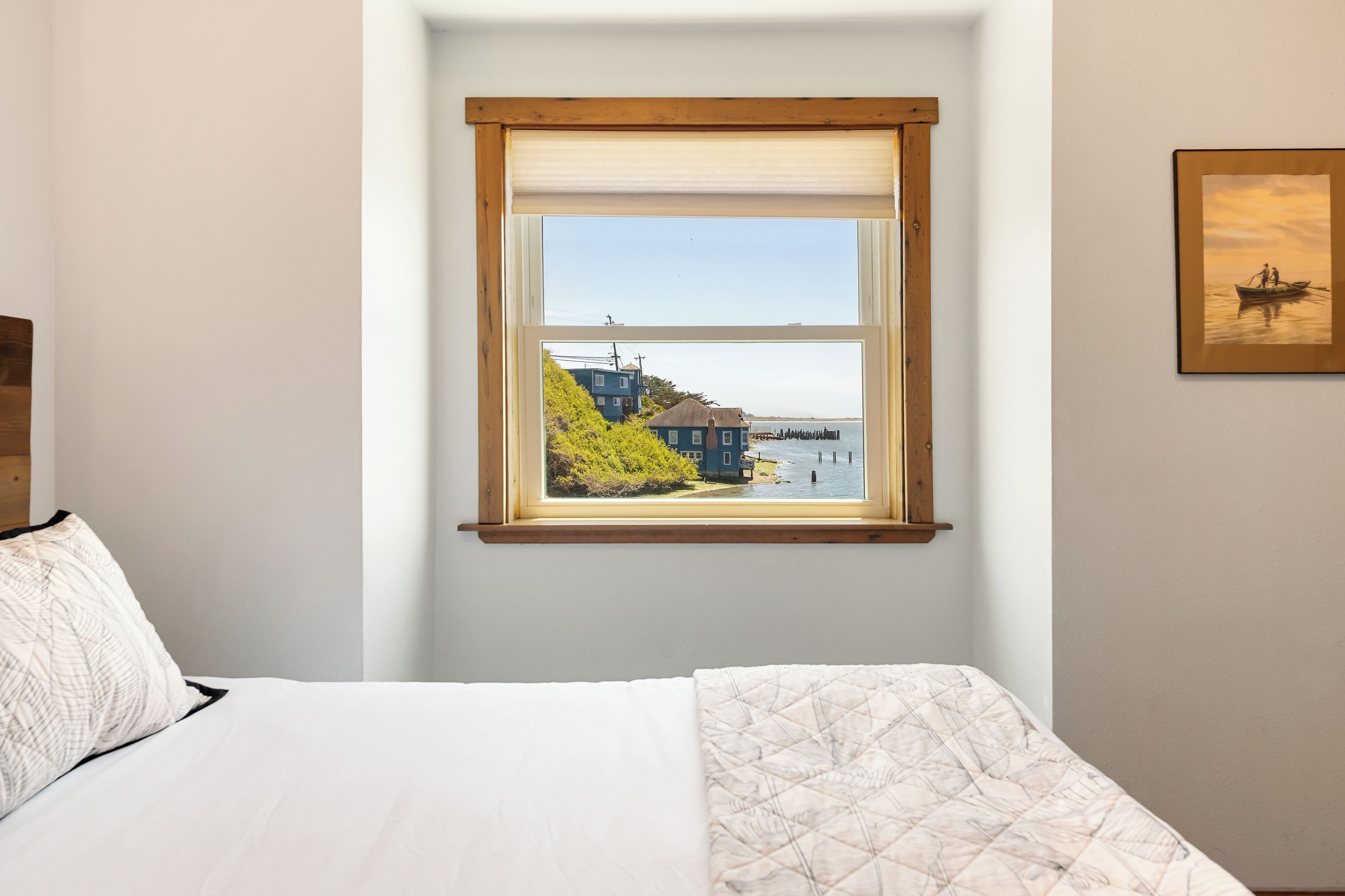 Ein Schlafzimmer mit einem Bett, einem Fenster mit Blick auf ein Haus und Wasser, und einem gerahmten Bild an der Wand.