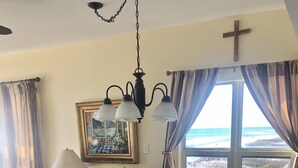 A dining area with a glass table, cushioned chairs, a chandelier, a painting, and a view of the beach.