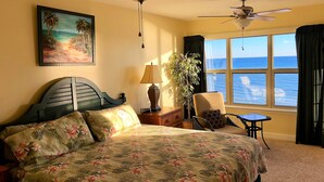A bedroom with a bed, a chair, a table, a lamp, a plant, and a painting of a beach scene.