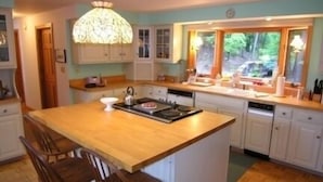 A kitchen with a central island, wooden chairs, a chandelier, and a window with a view of greenery.