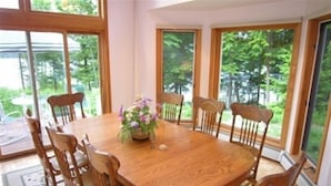 A round wooden dining table with six chairs, a floral centerpiece, and large windows offering a view of greenery.