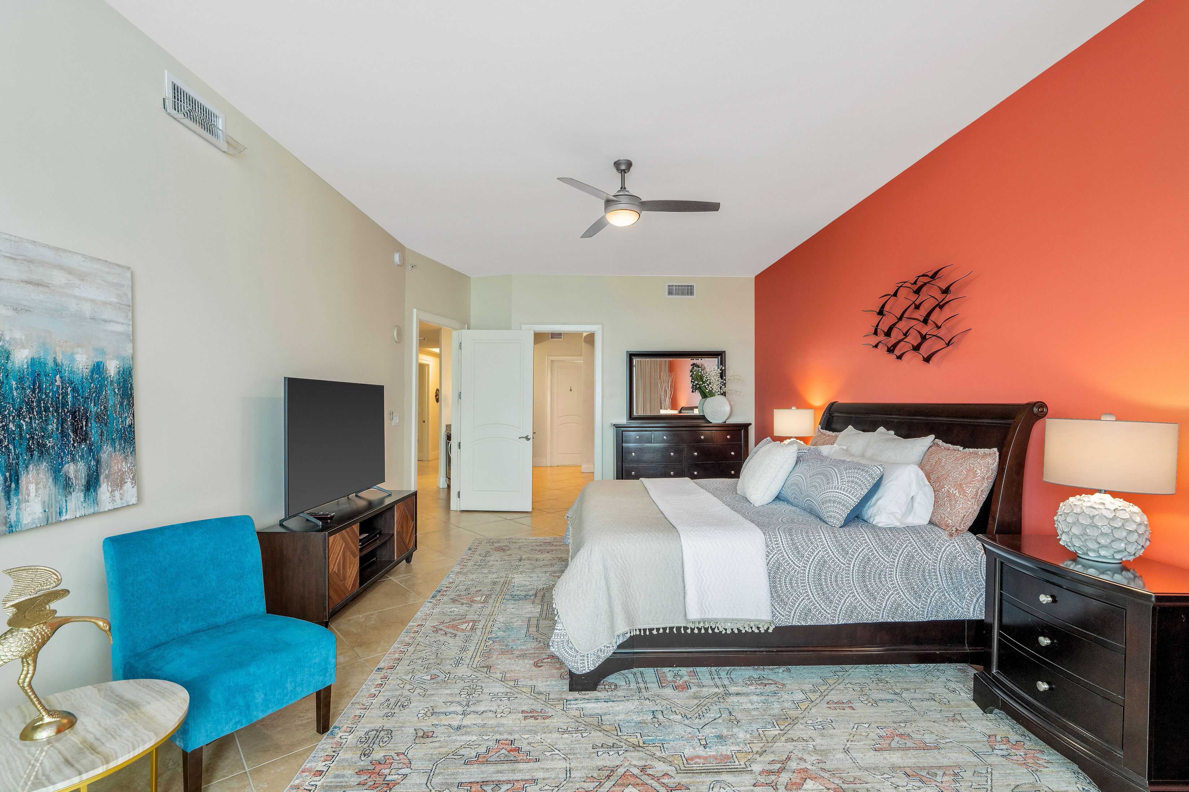A bedroom with a bed, a blue armchair, a TV, and a ceiling fan.