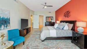 A bedroom with a bed, a blue armchair, a TV, and a ceiling fan.