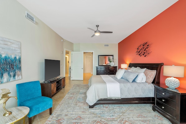 A bedroom with a bed, a blue armchair, a TV, and a ceiling fan.