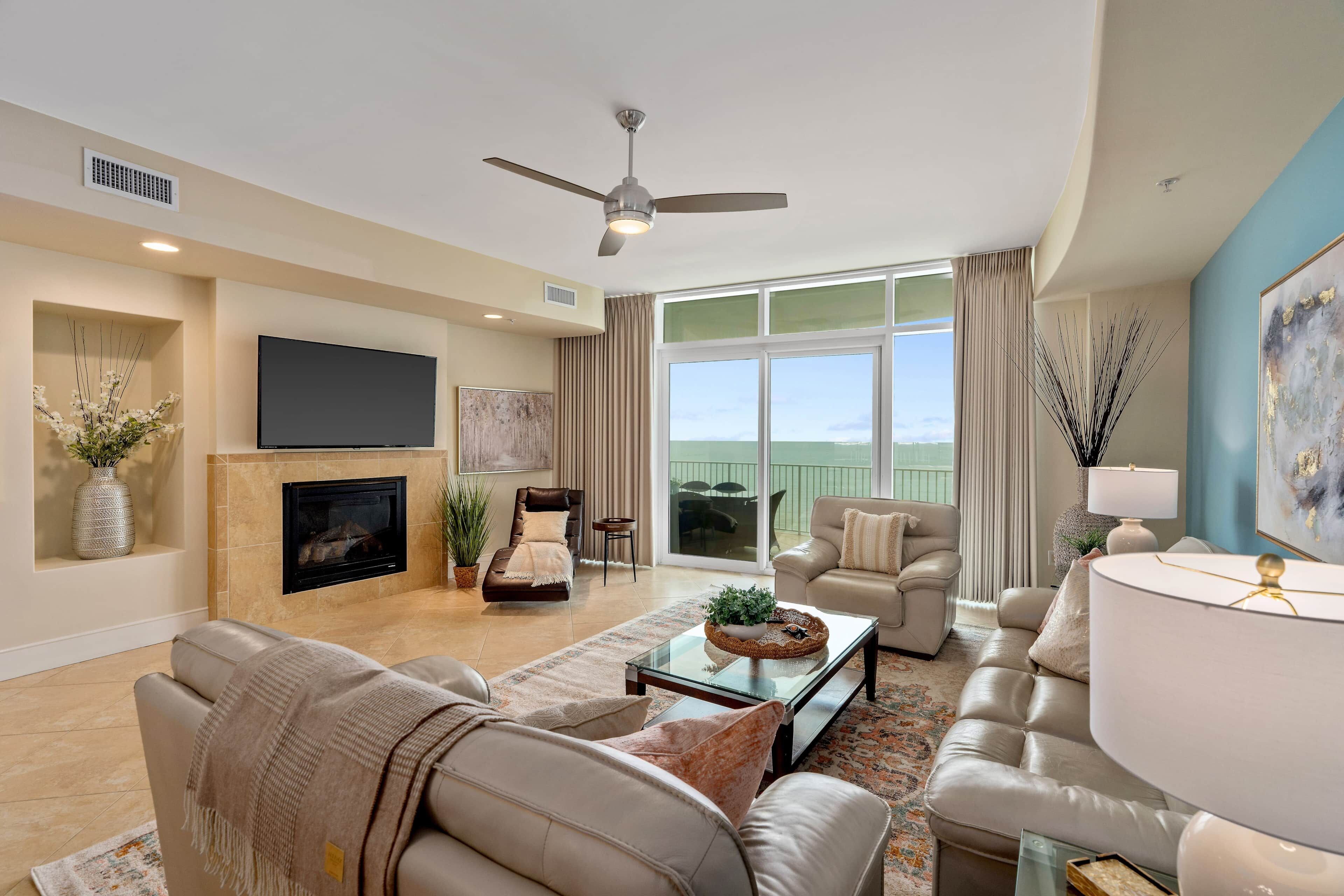 A spacious living room with a fireplace, comfortable seating, a flat-screen TV, and a view through large windows.