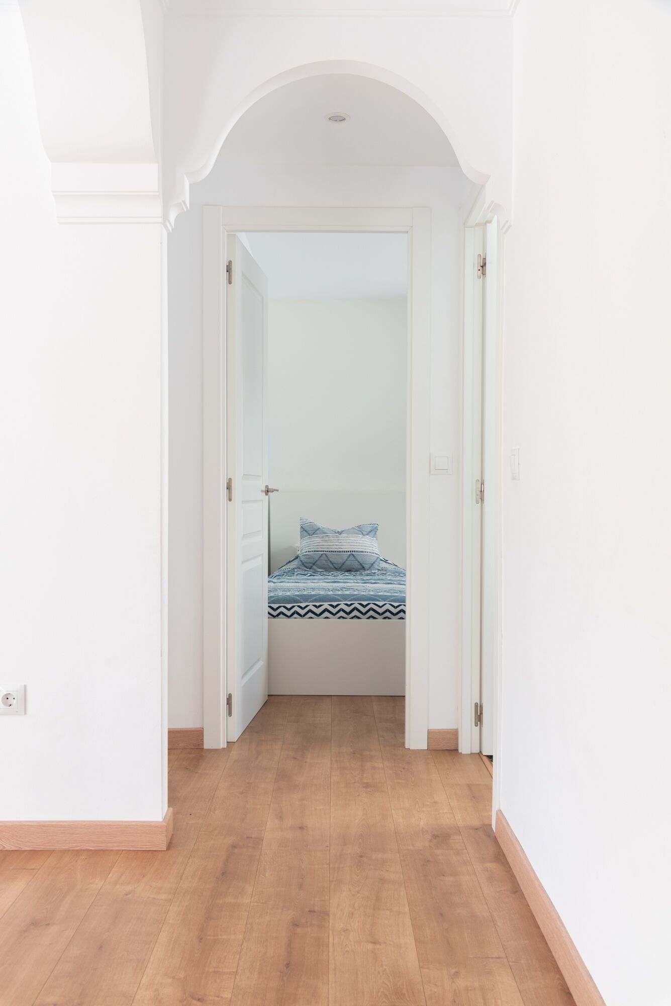 Ein schmaler Gang führt zu einem Schlafzimmer mit einem Bett, Holzboden und weißen Wänden.