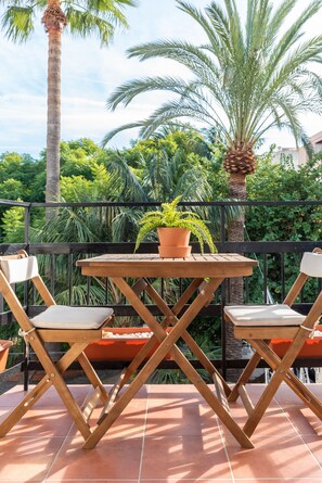 An outdoor patio with a wooden table and two chairs, surrounded by lush greenery and palm trees.