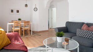 Een moderne woonkamer met een grijze bank, een glazen salontafel en een eethoek met houten stoelen en een witte tafel.