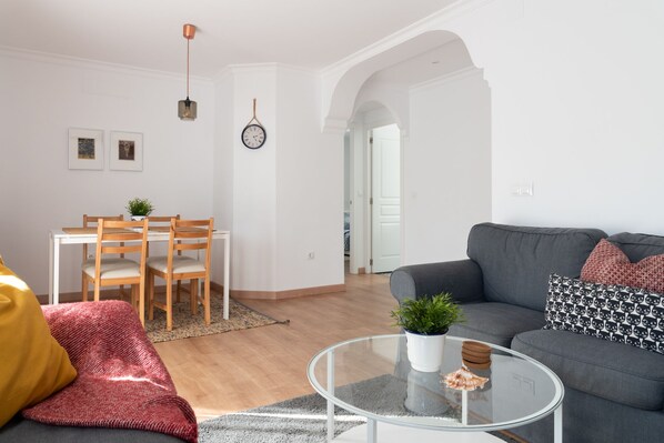 A modern living room with a grey sofa, a glass coffee table, and a dining area with wooden chairs and a white table.