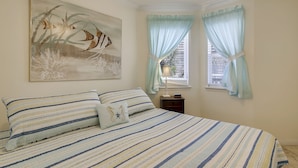 A bedroom with a bed, striped bedding, a wall-mounted painting, and two windows with light blue curtains. - Beach Cottage steps from Surf... fishing... the Beach and Serene Sunsets (1303) (Galveston)