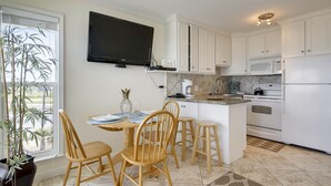 A kitchen with white cabinets, a flat-screen TV, a dining table with chairs, and a potted plant. - Beach Cottage steps from Surf... fishing... the Beach and Serene Sunsets (1303) (Galveston)