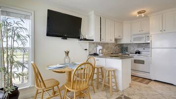 A modern kitchen with white cabinets, a flat-screen TV mounted above the counter, and a dining area with wooden chairs and a table.