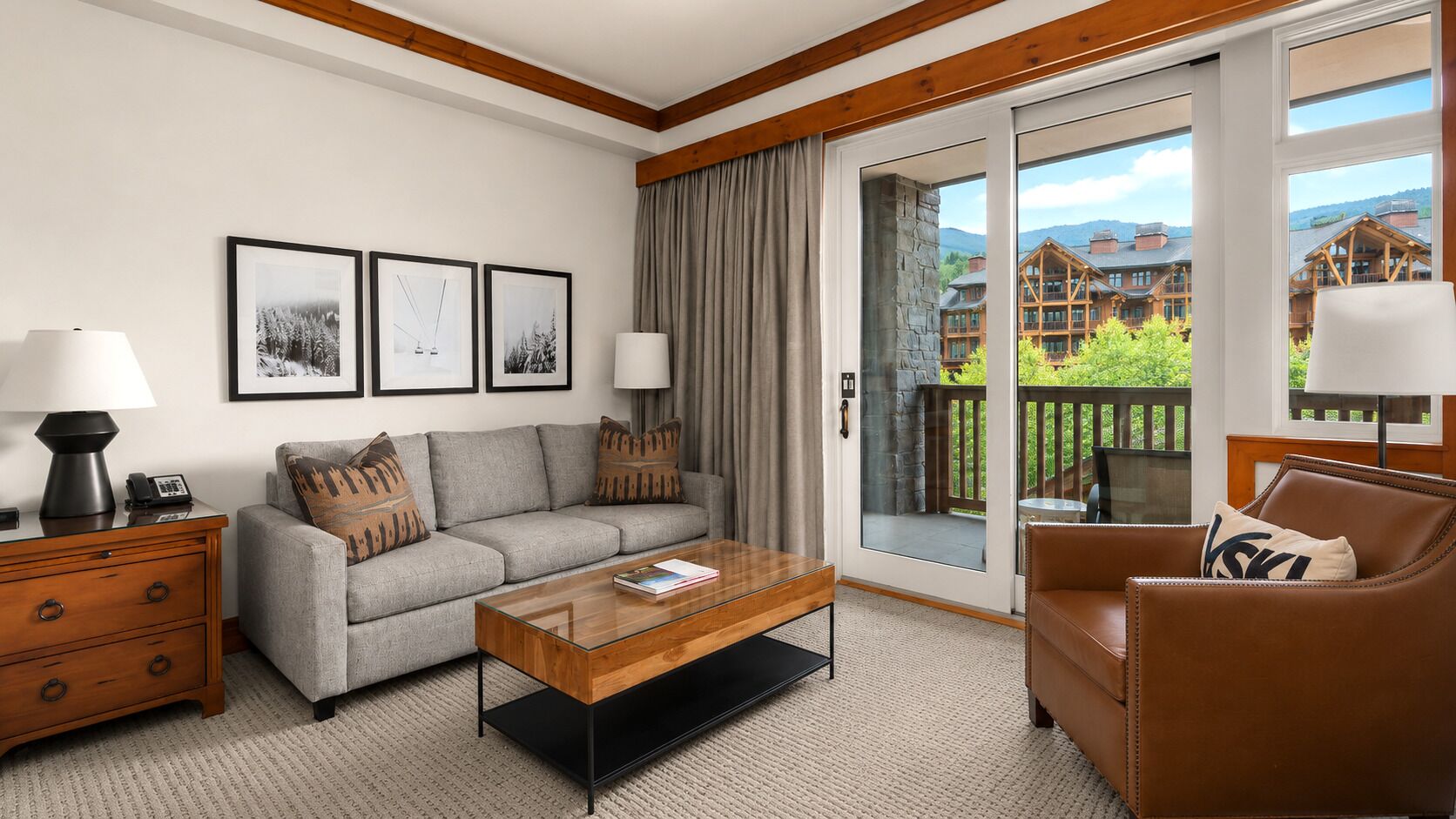 A modern hotel room with a sofa, a leather armchair, a wooden coffee table, and a view of a mountain landscape through the glass door.
