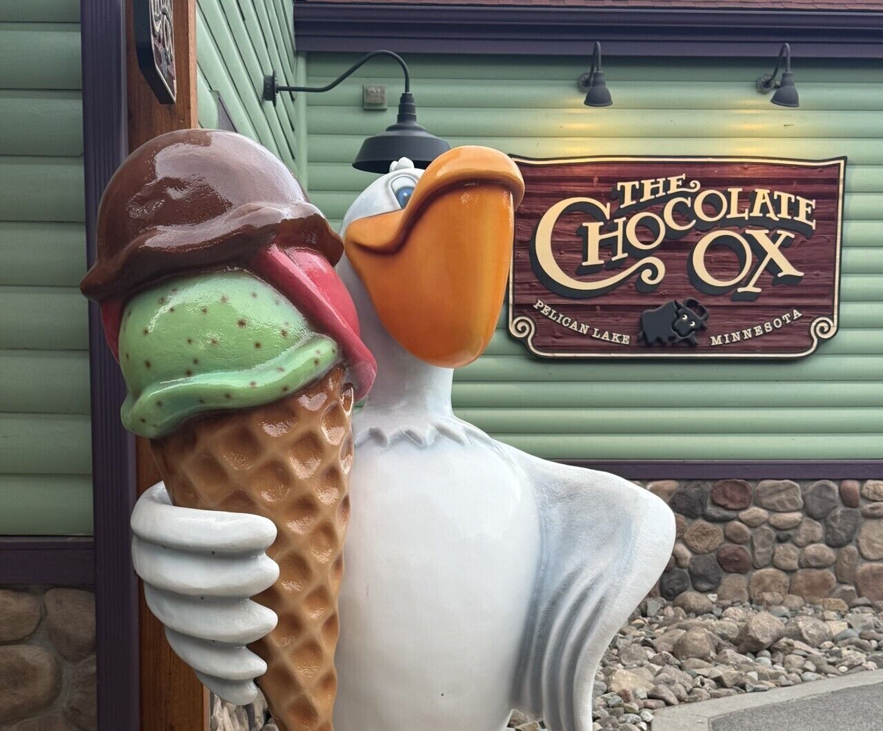A sculpture of a duck holding an ice cream cone in front of a building with a sign that reads 'The Chocolate Ox' in Pelican Lake, Minnesota.