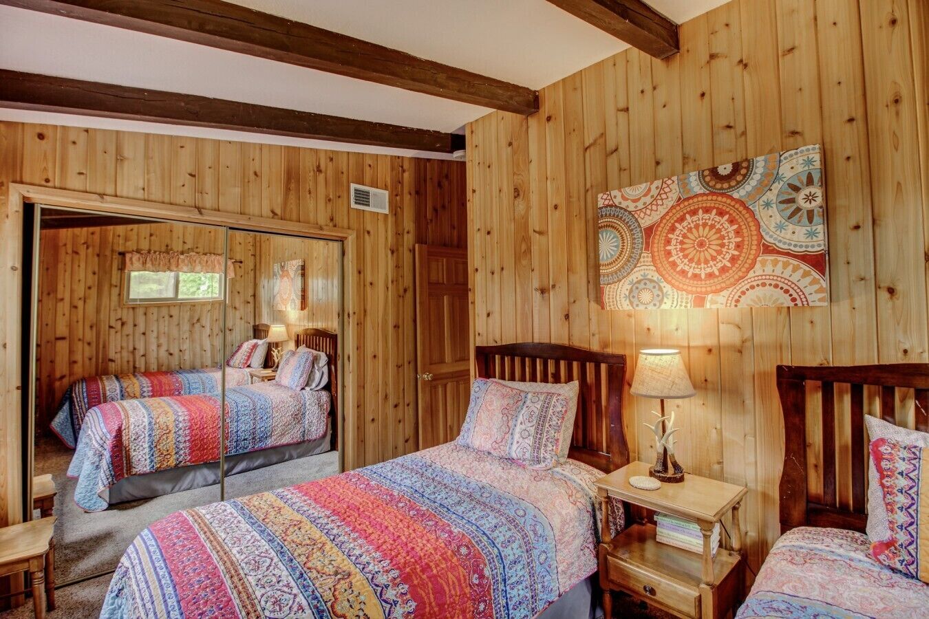 Una habitación con paredes de madera, dos camas con ropa de cama estampada, un espejo, un tapiz y una pequeña mesa con una lámpara.