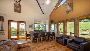 A spacious living room with a wooden floor, leather sofas, a bar area with stools, and large windows offering a view of the outdoors. - Incredible Mtn Views - 106 Acres: Private Pool, Pickleball Fall of 2026 (Zionville)