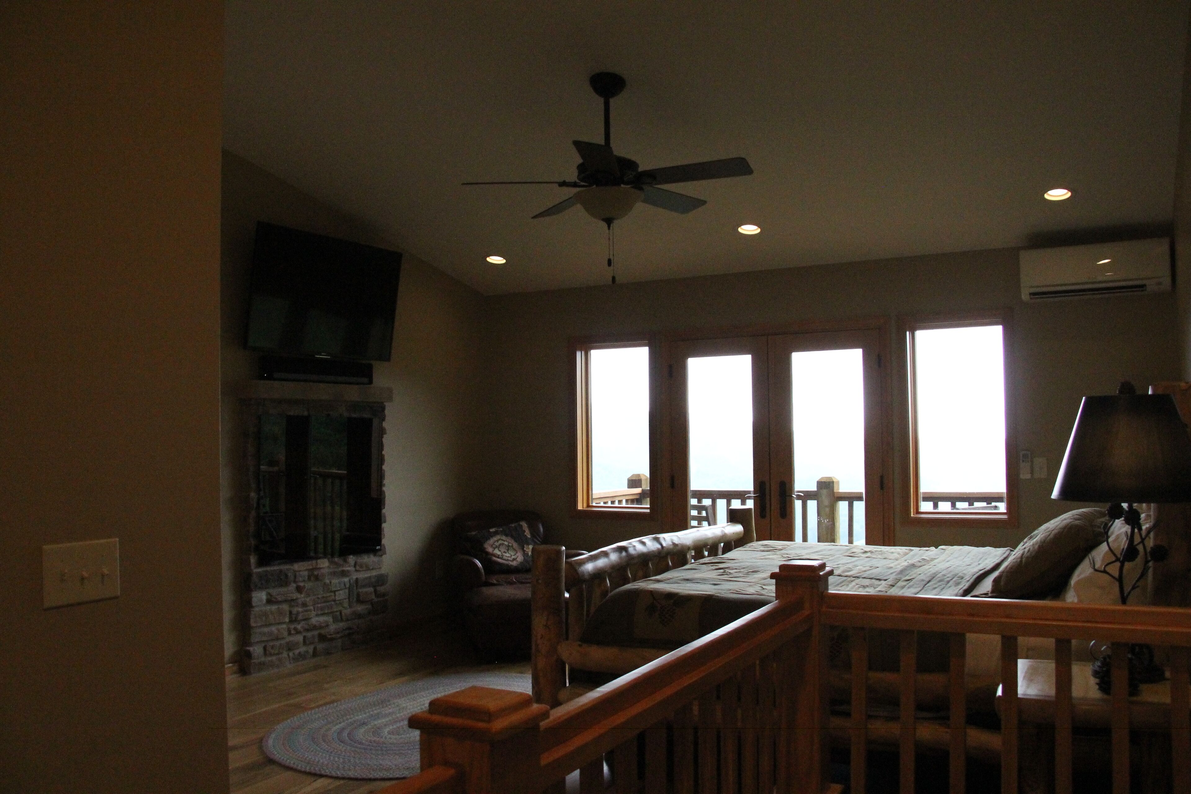 A bedroom with a bed, a wooden headboard, a ceiling fan, a stone fireplace, and a large window.