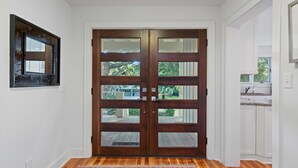 Una puerta doble de madera con paneles de vidrio, un tapete estampado y vistas a un patio trasero con terraza de madera y vegetación.