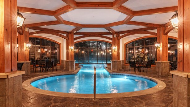 Une grande piscine intérieure dotée d’un plafond en bois et d’un sol en pierre, entourée d’espaces de détente et de grandes fenêtres.