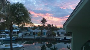 Un puerto deportivo con barcos amarrados, una masa de agua tranquila que refleja el cielo, y una zona residencial con casas y palmeras.