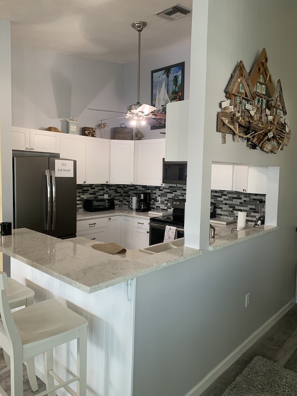 A modern kitchen with white cabinets, a marble countertop, and a built-in stove. There is a refrigerator with a sign indicating "151 3rd Street." A ceiling fan and a decorative wooden piece are also visible.