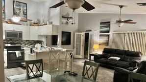 Una cocina moderna con armarios blancos, un microondas y una estufa. Hay un comedor con una mesa de vidrio y sillas. Un sofá de cuero negro se encuentra en la sala.