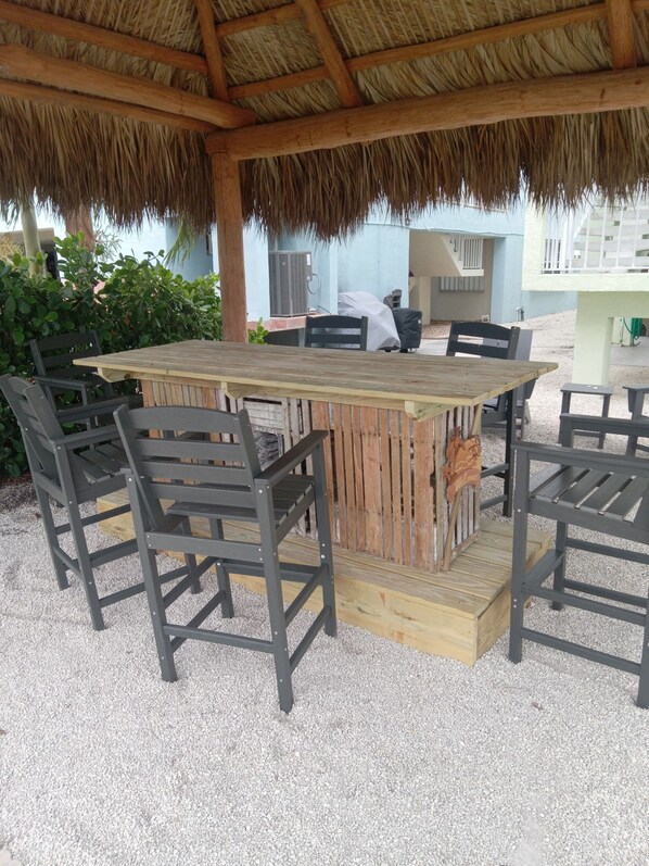 An outdoor dining area with a wooden table and chairs, a thatched roof, and a view of a building in the background. - Canal side location with beach & pool club membership! (Key Colony Beach)
