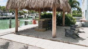 Un área de estar al aire libre con techo de paja, una mesa y sillas, un muelle con barcos y vistas al frente del agua.