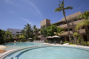 A swimming pool with lounge chairs and umbrellas, surrounded by palm trees and a multi-story building.