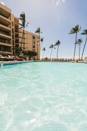 Piscina com água cristalina, rodeada por palmeiras e um edifício com várias varandas.