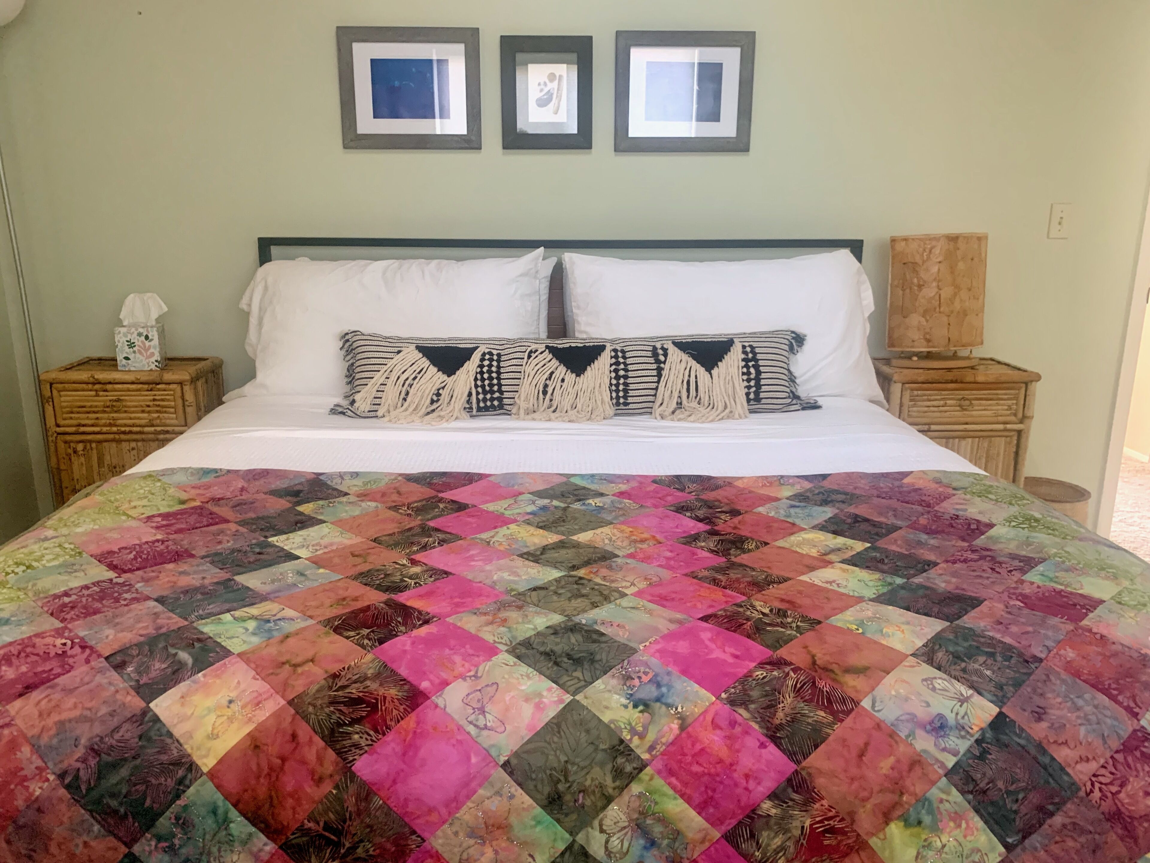 A bed with a colorful quilt, two framed pictures on the wall, and a wooden nightstand with a tissue box.