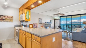 A spacious kitchen with wooden cabinets, a central island, and a large window overlooking the ocean. - Stunning Oceanfront Retreat, Kahana Reef 218  (Lahaina)