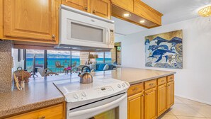 A modern kitchen with wooden cabinets, a microwave, stove, and a large window overlooking the ocean. - Stunning Oceanfront Retreat, Kahana Reef 218  (Lahaina)